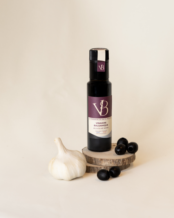 ail et olives 3 Bouteille de vinaigre balsamique ail et olive noire posée sur des rondins de bois, accompagnée d’une gousse d’ail et de plusieurs olives noires