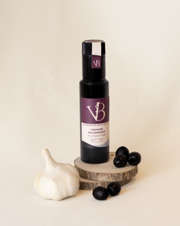 ail et olives 4 Bouteille de vinaigre balsamique ail et olive noire posée sur des rondins de bois, accompagnée d’une gousse d’ail et de plusieurs olives noires