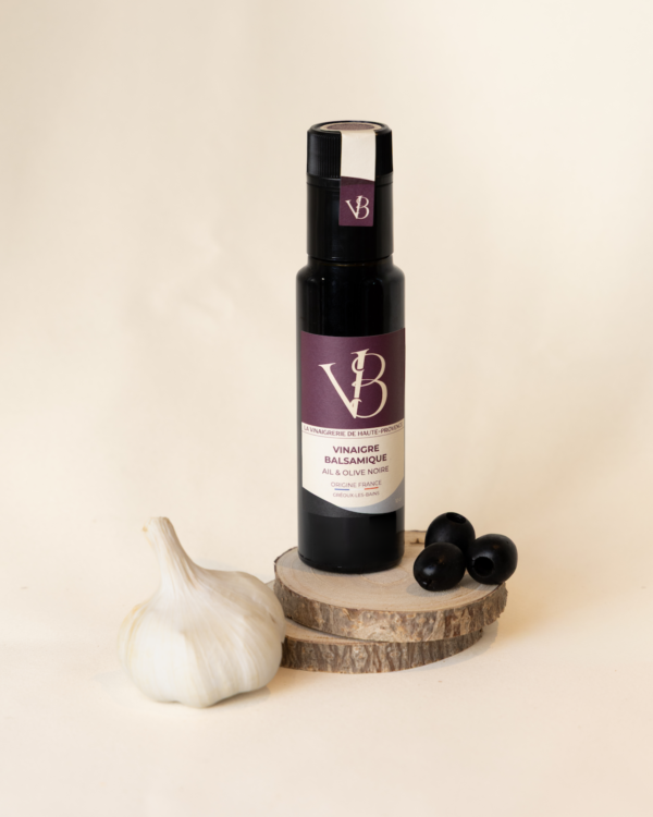 ail et olives 5 Bouteille de vinaigre balsamique ail et olive noire posée sur des rondins de bois, accompagnée d’une gousse d’ail et de plusieurs olives noires