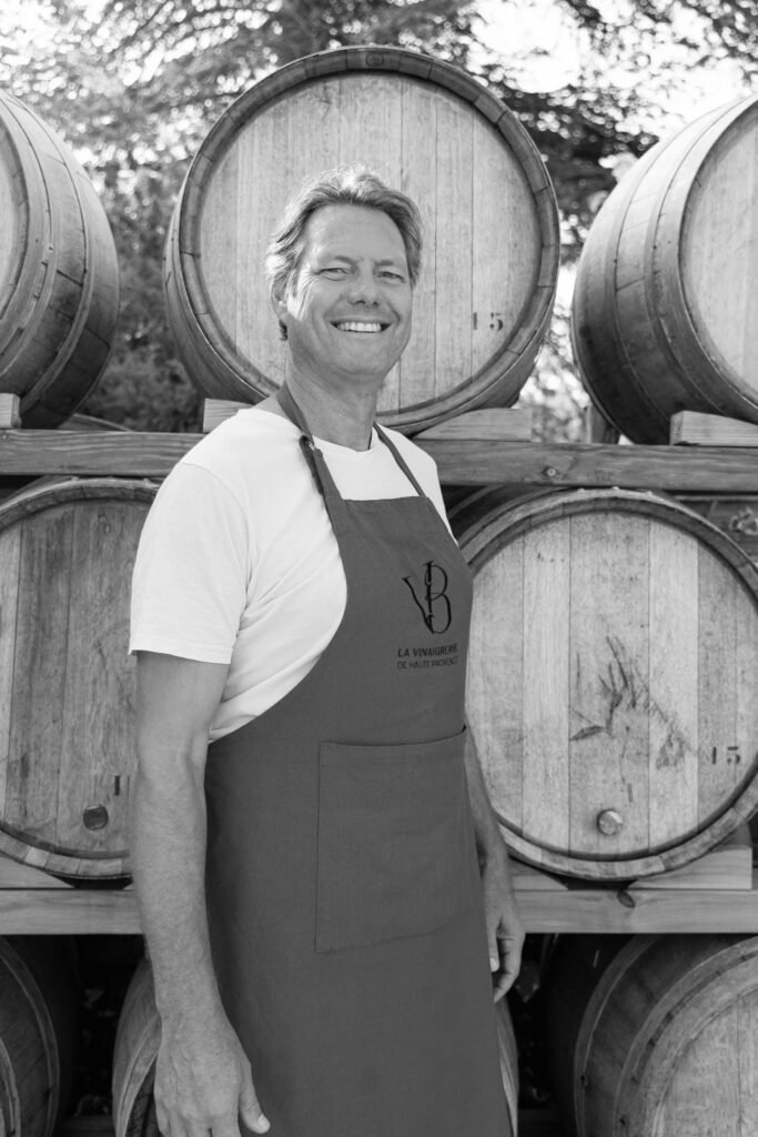 Portrait en noir et blanc d’un maître vinaigrier souriant devant des fûts en bois, portant un tablier marqué "La Vinaigrerie de Haute-Provence"